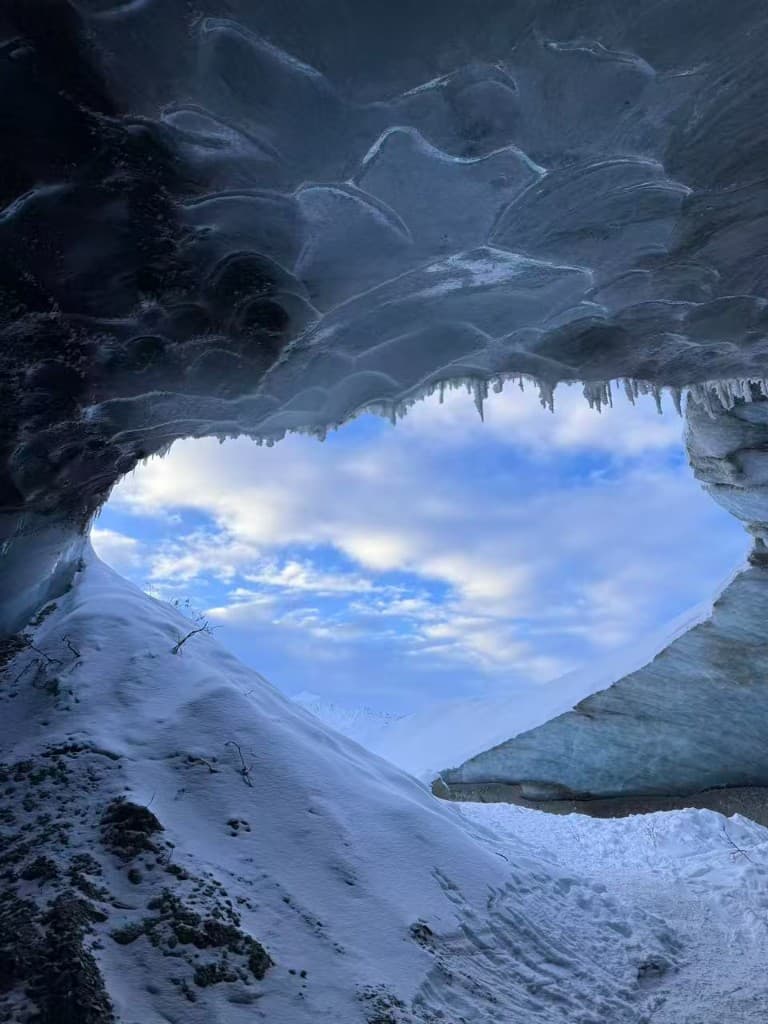 View from inside an ice cave toward snowy peaks and blue sky, Castner Glacier, Alaska